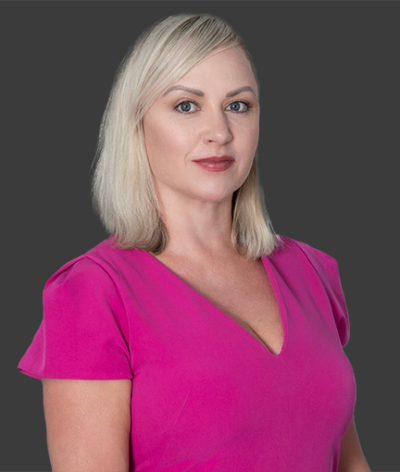 Katya Gozias in a pink shirt is standing in front of a gray background